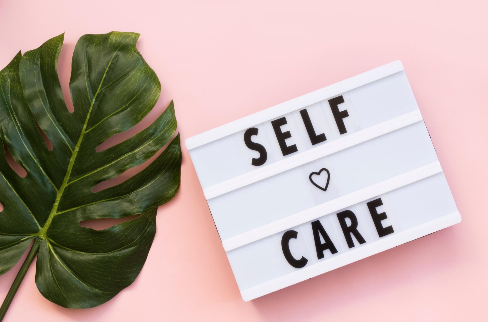 A sign that says self care on a pink background next to a leaf